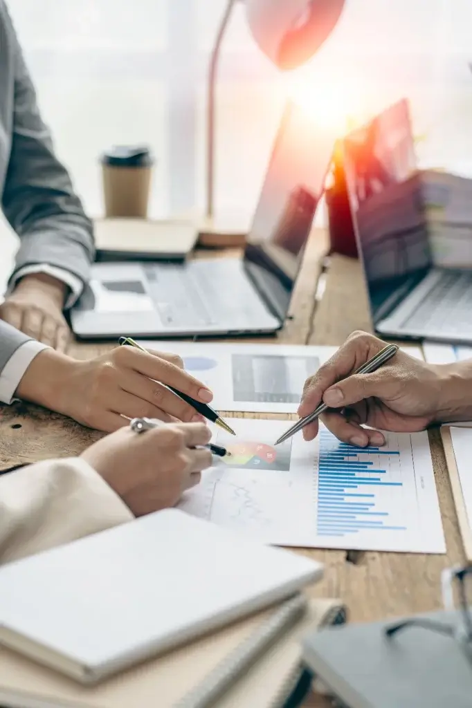 Close-up of hands pointing at business charts and graphs on desk with laptops and coffee cup in background